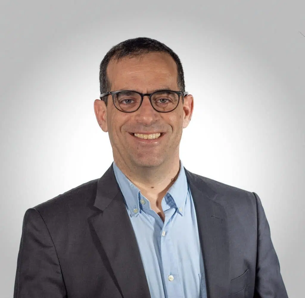 A man wearing glasses, a blue button-up shirt, and a dark blazer is smiling in front of a plain light background.
