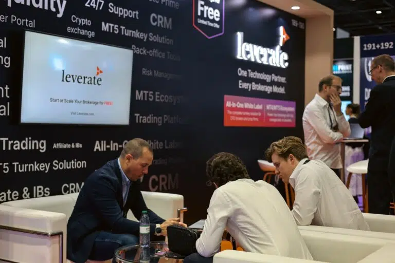 Three men sit and talk at a booth for Leverate, a financial technology company, at a business expo. A large screen and marketing banners are visible in the background.
