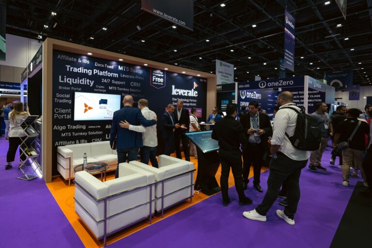 Un concurrido recinto ferial con gente interactuando en un stand que promociona plataformas comerciales, rodeado de expositores, sillas y señalización de marca.
