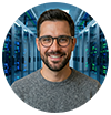 Un homme aux cheveux noirs, à la barbe et aux lunettes sourit alors qu'il se tient dans une salle de serveurs avec des rangées de serveurs informatiques à l'arrière-plan.